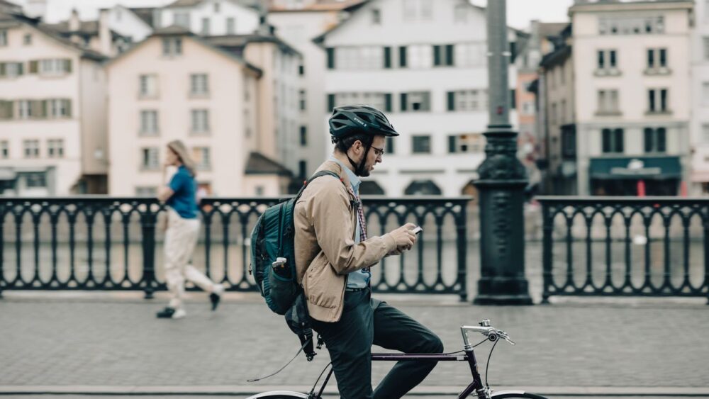 A male bicycle commuter wearing a helmet, beige jacket, and backpack sits astride his bike on a bridge, looking down at his smartphone with historic European buildings in the background.