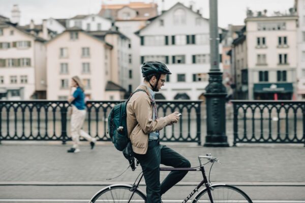 A male bicycle commuter wearing a helmet, beige jacket, and backpack sits astride his bike on a bridge, looking down at his smartphone with historic European buildings in the background.