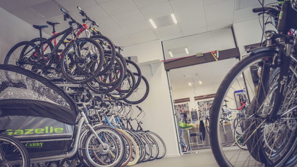 A wide-angle view of a bicycle shop interior, featuring rows of mountain and city bikes organized on floor stands and multi-tiered wall racks, with a "Gazelle" brand child carrier bicycle in the foreground.