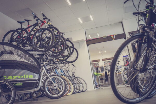 A wide-angle view of a bicycle shop interior, featuring rows of mountain and city bikes organized on floor stands and multi-tiered wall racks, with a "Gazelle" brand child carrier bicycle in the foreground.