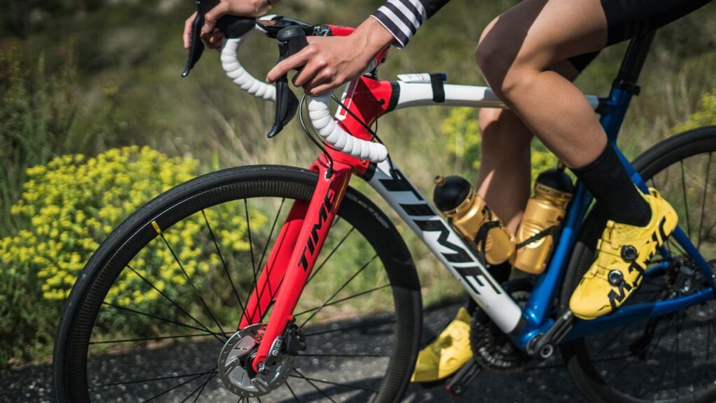A close-up, dynamic shot of a cyclist in action, riding a white, red, and blue "Time" brand carbon road bike. The cyclist is wearing bright yellow cycling shoes and gripping the white-wrapped drop handlebars on an outdoor road.