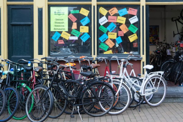 A street-level view of a bicycle shop storefront with a row of city bikes parked outside. The windows are decorated with colorful paper signs, and a main sign reads "Inkoop, Verkoop, Verhuur, Reparatie."