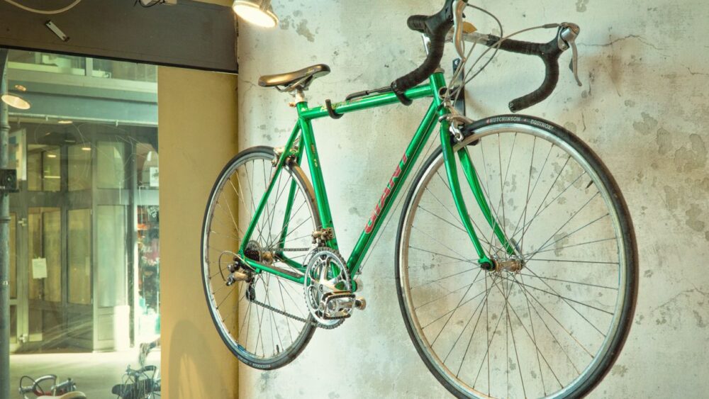 A bright green vintage road bicycle mounted horizontally as a display on a textured, light-colored concrete wall under an overhead spotlight.
