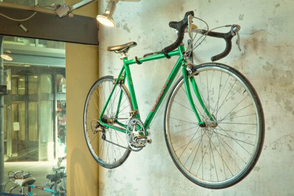 A bright green vintage road bicycle mounted horizontally as a display on a textured, light-colored concrete wall under an overhead spotlight.