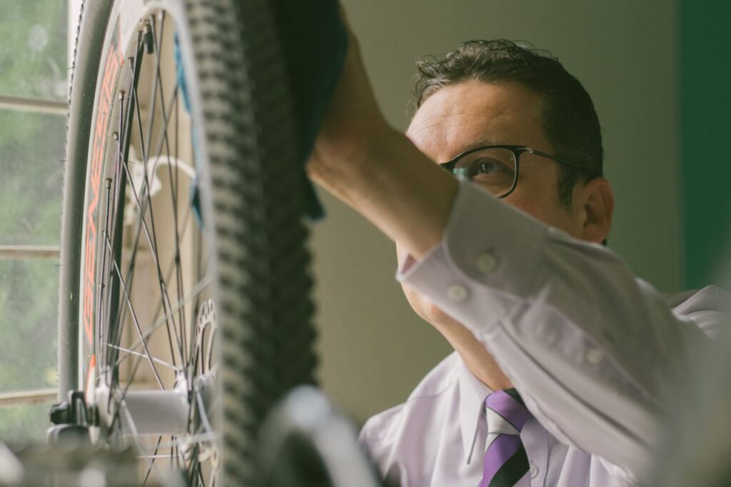 A close-up shot of a man wearing glasses, a white dress shirt, and a purple tie using a cloth to wipe down the front tire of a mountain bike indoors.