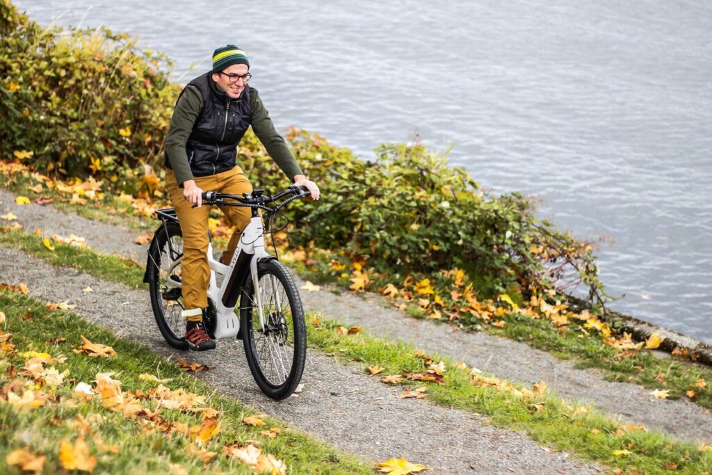 A man smiling while riding a white electric bicycle on a gravel path next to water in the autumn.