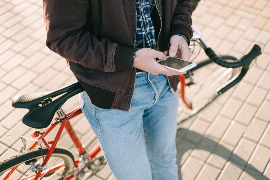 A close-up, mid-section shot of a person in a brown jacket and light-blue jeans leaning against a red bicycle while using a white smartphone.