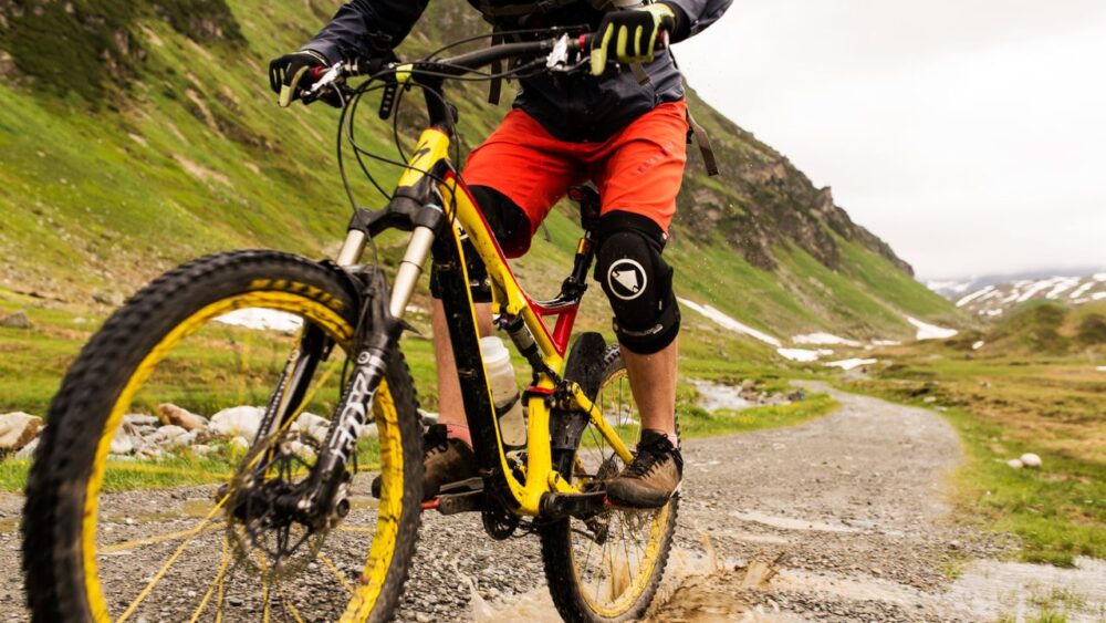A cyclist riding a yellow mountain bike through a water puddle on a mountain trail.