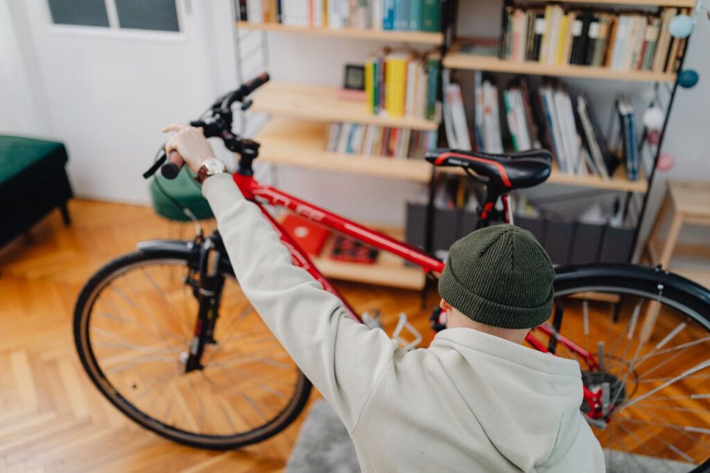 An over-the-shoulder view of a person wearing a green knit beanie and a light grey hoodie holding the handlebars of a red bicycle in a cozy apartment with a packed bookshelf in the background.