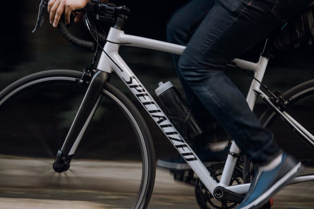 A close-up, side-profile view of a person in dark jeans pedaling a white Specialized road bike, showing motion blur on the wheels and feet.