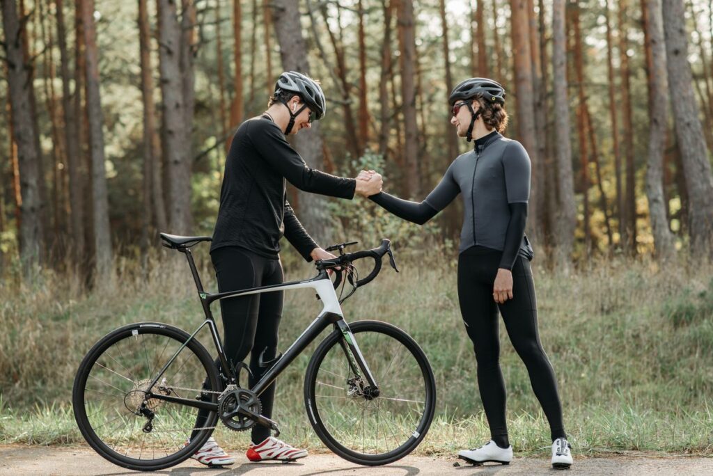 Two cyclists in helmets and dark athletic cycling gear sharing a friendly handshake next to a black and white road bicycle on a paved path lined with tall pine trees.