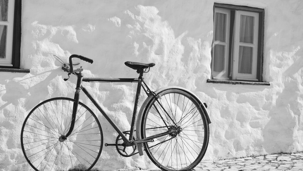 A black and white photograph of a classic vintage bicycle leaning against a textured white stucco wall next to a small window.