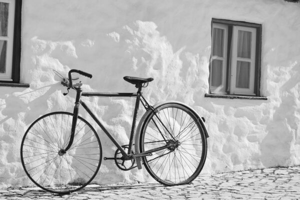 A black and white photograph of a classic vintage bicycle leaning against a textured white stucco wall next to a small window.